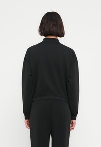 Μαύρο φούτερ με ψηλό γιακά, κατεβασμένους ώμους και ριμπάτο τελείωμα. Ομαλή υφή χωρίς εμφανή μοτίβα ή μεταλλικά διακοσμητικά.