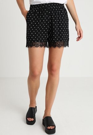 Femme portant un short noir à pois avec un bord en dentelle et des sandales plateformes noires, debout devant un fond uni.