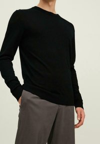 Pull noir à manches longues, col rond, poignets et ourlet côtelés, associé à un pantalon gris. Texture tricotée, coupe près du corps.