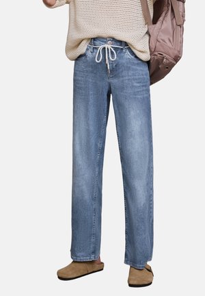 Person trägt hellblaue Jeans mit weitem Bein und weißem Kordelzug, beigen Stricktop und braune Slipper, und hat einen mauvefarbenen Rucksack dabei.