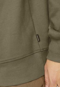 Olívazöld pulóver, bordás mandzsettával és szegéllyel, puha tapintású, a varrás mellett egy kis fekete márkajelzés látható.