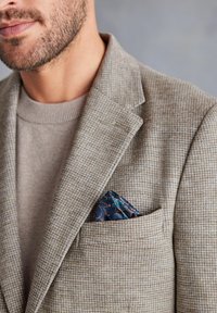 Brauner Hahnentritt-Blazer mit Reverskragen; grauer Pullover darunter; blauer gemusterter Einstecktuch in der Brusttasche.