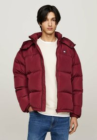 Jachetă puffer maroon cu închidere cu fermoar, glugă și design matlasat orizontal. Prezintă o etichetă cu logo pe piept. Purtați-o peste un pulover ușor.