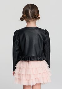Czarna kurtka z sztucznej skóry z bufiastymi rękawami i falbaniastym dołem, noszona na jasnoróżowej, warstwowej spódnicy z tiulu, widziana z tyłu.
