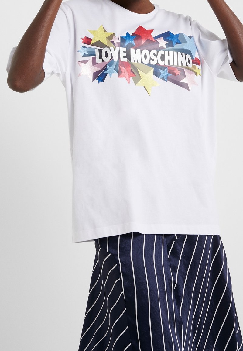 Bílý bavlněný tričko s multibarevným grafickým vzorem hvězdy a odvážným textem "LOVE MOSCHINO." Modelka má na sobě pruhovanou námořnickou sukni.