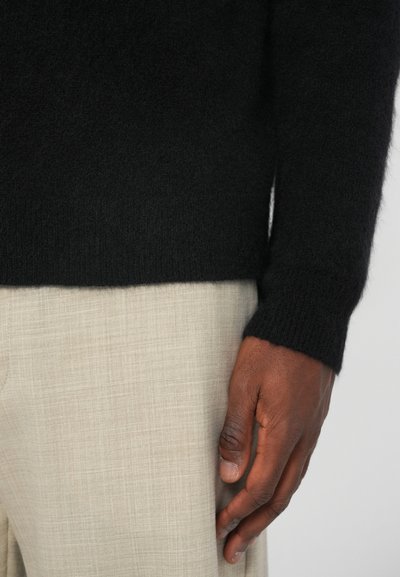 Maglione di cashmere nero con una texture morbida e maniche aderenti, indossato con pantaloni in lino beige chiaro. Primo piano di una mano appoggiata sui pantaloni.