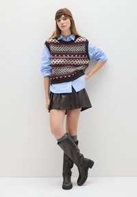 Strickweste mit geometrischen Mustern in erdigen Tönen, über einem hellblauen Hemd getragen. Brauer Lederfaltenrock und hohe braune Stiefel.