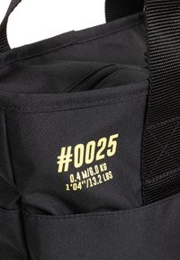 Musta kangaslaukku, jossa on teksturoitu pinta, vetoketjullinen tasku ja keltainen painettu etiketti, jossa on mitat ja paino.