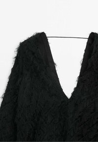 Vêtement en tissu texturé noir avec un décolleté en V profond suspendu à un cintre noir fin sur un fond blanc.