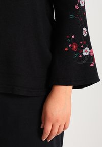 Haut noir à manches évasées avec broderie florale colorée en rouge, rose et vert sur la manche. Tissu doux avec une coupe ample.