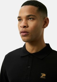 Czarna koszulka polo z kołnierzykiem, z guzikiem i kwadratową łatą logo w kolorze złotym na lewej piersi. Gładka tekstura materiału.