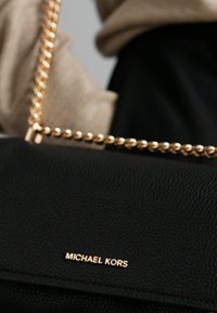 Zwarte leren handtas met een reliëfstructuur, voorzien van een gouden kettingriem en een metalen Michael Kors-logo aan de voorkant.