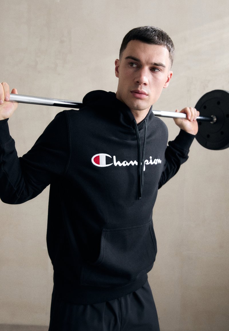 Haut à capuche noir avec une poche frontale et un logo comportant du texte rouge et blanc. Le modèle tient une barre, affichant un look décontracté et athlétique.
