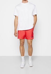 Muž oblečený v jednoduchém bílém tričku s krátkým rukávem, jasně červených šortkách Nike, bílých ponožkách a bílých teniskách, stojící na světlém pozadí.