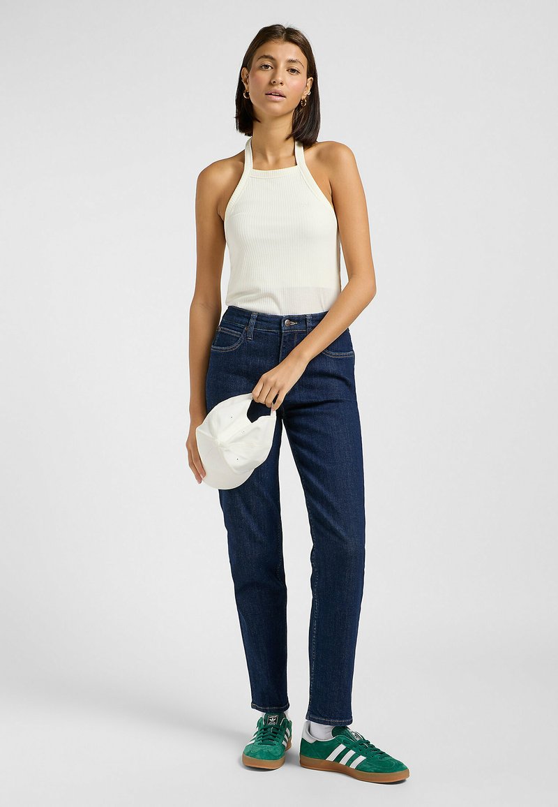 Biały, żeberkowany top na halter, ciemnoniebieskie dżinsy, zielone trampki z białymi paskami i biała czapka trzymana w ręku. Gładka tekstura i dopasowany krój.