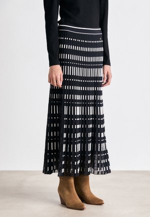 Femme portant une jupe midi plissée noire et blanche à motif, associée à un haut noir à manches longues et des bottines en daim marron.