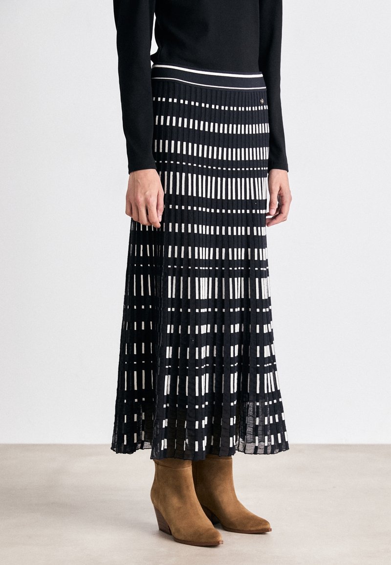 Femme portant une jupe midi plissée noire et blanche à motif, associée à un haut noir à manches longues et des bottines en daim marron.