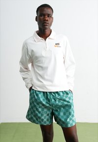 Hvit langermet polo med glidelås og Nike-logo, paired med grønne rutete shorts, som viser en avslappet passform og teksturert stoff.