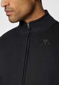 Casaco preto com fecho zip, com colarinho alto, feito de material suave. Apresenta um logo em tom sobre o peito e um detalhe de fecho metálico.