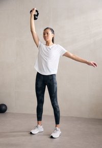 Juodas ketlebellas laikomas virš galvos žmogaus, kuris vilki baltą trumpą rankovę marškinėlį ir juodas kamufliažines leggingus, derinamus su baltais sportiniais batais.
