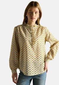 Bluza cu mâneci lungi, realizată dintr-un material ușor, cu fundal alb, decorată cu modele florale galbene și cu un guler cu volane.