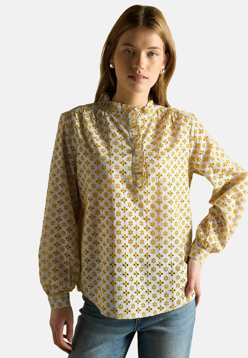 Bluza cu mâneci lungi, realizată dintr-un material ușor, cu fundal alb, decorată cu modele florale galbene și cu un guler cu volane.