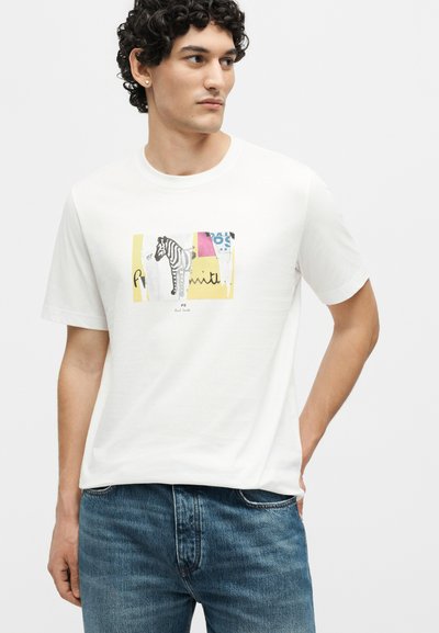 Άνδρας με σγουρά μαλλιά που φοράει λευκό μπλουζάκι με σχέδιο ζέβρας και πολύχρωμους κύβους, συνδυασμένο με μπλε τζιν, σε απλό φόντο.