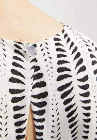 Valkoinen kangas mustalla lehtikuviolla, jossa on nappisulku takana ja avonainen venttiili kauluksessa. Sujuva tekstuuri.