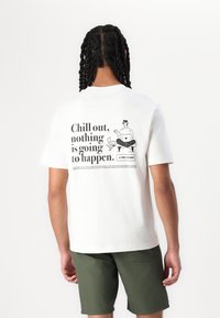 Weißes Baumwoll-T-Shirt mit grafischem Design auf der Rückseite, das schwarzen Text und eine Illustration zeigt. Kurzärmelig und lässige Passform. Kombiniert mit olivfarbenen Shorts.