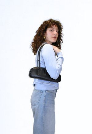 Μαύρη δερμάτινη τσάντα χειρός με στρογγυλεμένο σχήμα και διπλούς ιμάντες, κρατημένη από ένα άτομο που φοράει ανοιχτό μπλε πουλόβερ και ελαφριά τζιν.