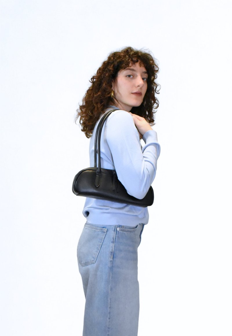 Μαύρη δερμάτινη τσάντα χειρός με στρογγυλεμένο σχήμα και διπλούς ιμάντες, κρατημένη από ένα άτομο που φοράει ανοιχτό μπλε πουλόβερ και ελαφριά τζιν.