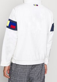 Chaqueta blanca con detalles en azul marino, que presenta parches coloridos en las mangas y la parte superior de la espalda. Textura suave, diseño casual con puños elásticos.