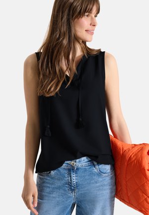 Kvinde iført sort top uden ærmer og blå jeans, som holder en orange vatteret jakke mod en ensartet lys baggrund.