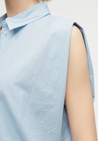 Αμάνικο φωτεινό μπλε πουκάμισο με λεπτές κάθετες λευκές ρίγες και μυτερό γιακά, φορεμένο από άτομο με ανοιχτό τόνο δέρματος.