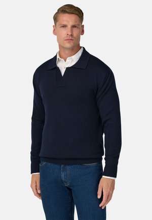 Jersey azul marino con textura de canalé, presenta un cuello ancho y una media tapeta. Combinado con una camisa blanca de cuello debajo.