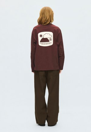 Personne aux cheveux mi-longs porte un t-shirt bordeaux à manches longues avec un logo blanc et un texte au dos, ainsi qu'un pantalon ample marron foncé.
