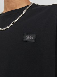 Crna pamučna majica s kvadratnom crnom etiketom na kojoj piše "JACK&JONES," uparena sa srebrnim ogrlicom.