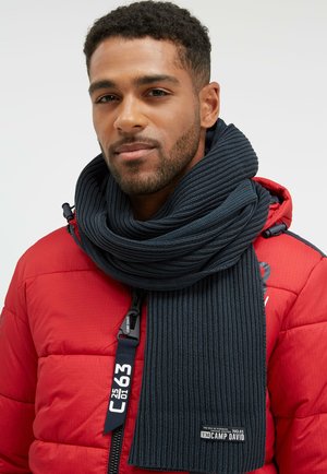 Dark navy knit scarf wrapped around the neck, featuring ribbed texture and a label, paired with a red puffer jacket.
