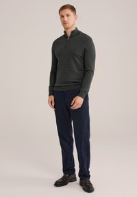 Fiatal férfi áll cipzáras, sötétszürke pulóverben, sötétkék nadrágban és fekete bőrcipőben egy sima háttér előtt.