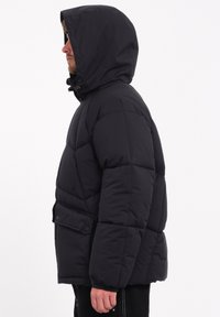 Zwarte pufferjack met capuchon, voorzien van een gestikte ontwerp, drukknoopjes op de voorkant van de zak en elastische manchetten voor warmte en comfort.