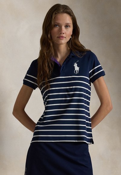 Jonge vrouw met lang haar, gekleed in een marineblauw poloshirt met witte horizontale strepen en een geborduurd polo-spelerlogo.