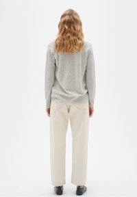 Femme aux cheveux blonds ondulés portant un pull gris clair, un pantalon blanc cassé et des chaussures noires, debout dos à un fond uni.