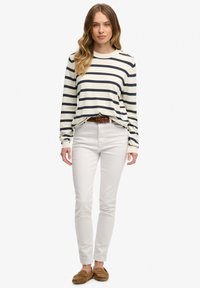 Kremowa bluzka w paski z długim rękawem w granatowe paski, zestawiona z białymi dopasowanymi dżinsami i brązowymi mokasynami. Delikatna faktura widoczna.