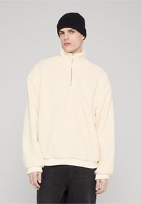 Cream-colored fleece pullover with a half-zip neckline, relaxed sleeves, and ribbed cuffs, paired with a black knitted beanie.