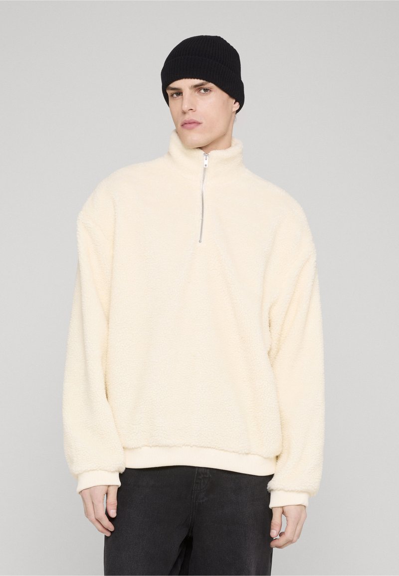Cream-colored fleece pullover with a half-zip neckline, relaxed sleeves, and ribbed cuffs, paired with a black knitted beanie.