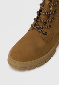 Botte de cheville en daim marron avec un embout arrondi, semelle en caoutchouc texturée et lacets tressés noirs et marron. Comprend des œillets métalliques.