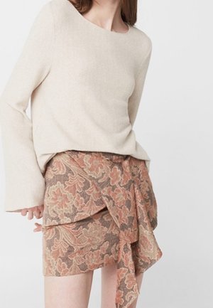 Femme portant un pull beige à manches longues et une jupe portefeuille taille haute à motif floral dans des tons de marron et rose.