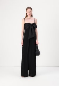 Combinaison noire avec un détail en nœud au niveau du buste, fines bretelles, jambes larges, en tissu lisse. La mannequin tient un petit sac à main noir.
