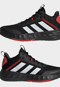 Zapatillas deportivas negras con acentos rojos; parte superior de malla texturizada, suela de goma, diseño de tres rayas blancas en los lados y tirador en el talón.