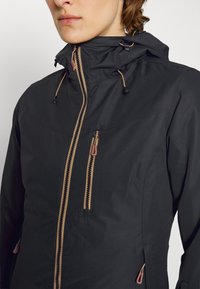 Schwarze wasserdichte Jacke mit hohem Kragen, kontrastierenden goldenen Reißverschlüssen, verstellbaren Kordeln und Reißverschlusstaschen auf der Vorderseite.